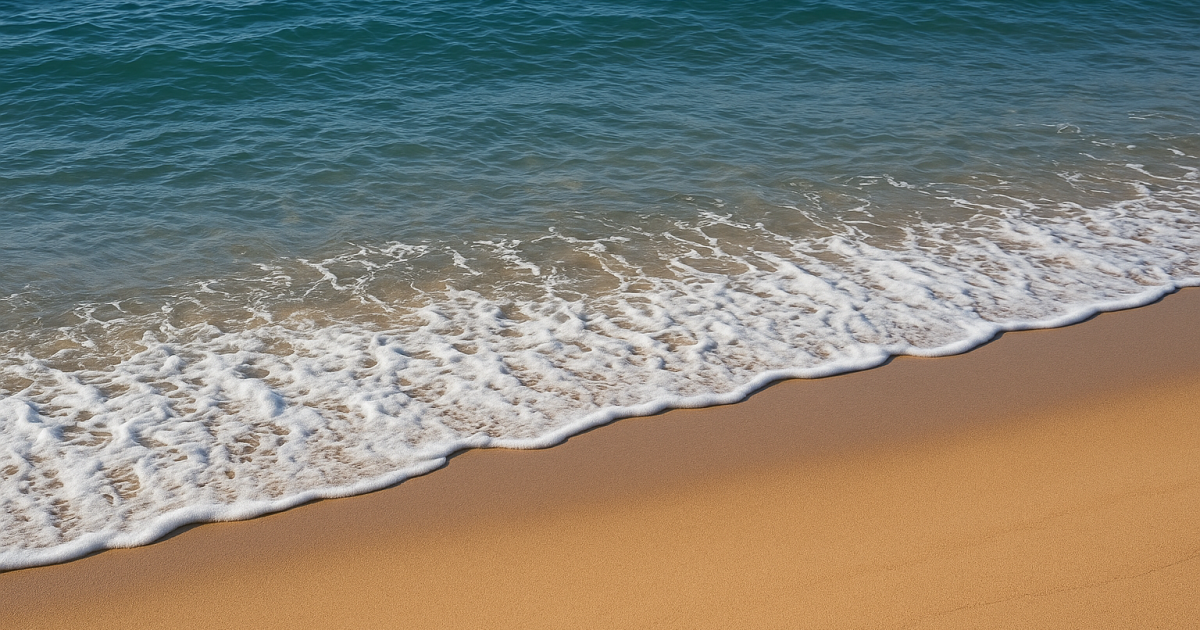 A gentle ocean wave washes onto a smooth sandy beach under clear daylight, with blue-green water meeting golden sand in a calm and peaceful scene.
